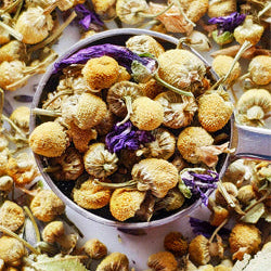 Dried herbs and flowers in a teaspoon on a bed of more dried herbs and flowers.