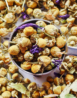 Dried herbs and flowers in a teaspoon on a bed of more dried herbs and flowers.